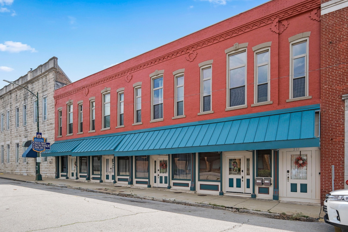 The historic storefront building features a vibrant red facade complemented by large windows. A blue awning extends over the entrance areas, which are decorated with holiday wreaths. A peaceful street scene is visible in front, showcasing the building's inviting atmosphere.