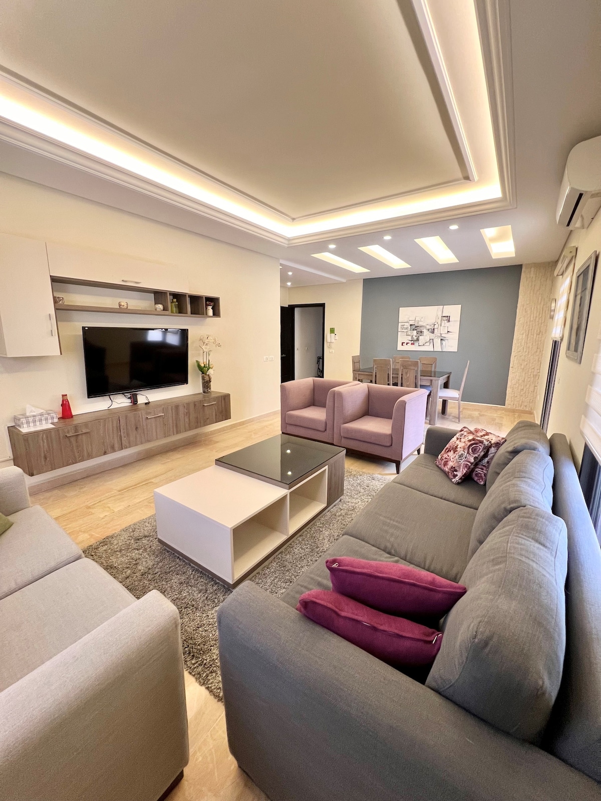 The living area is designed with modern furnishings, featuring a light gray sofa with purple accent pillows, a neutral rug, and a sleek coffee table. A wall-mounted television is prominently displayed, while a dining area with a wooden table is visible in the background.