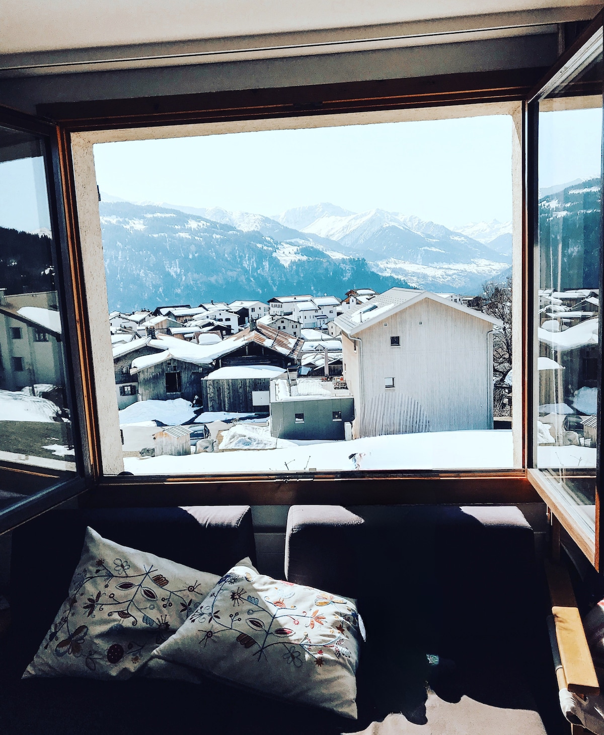 A panoramic view showcases the snow-covered rooftops of Falera along with the distant Swiss Alps. Bright natural light floods through the large open window, illuminating the cozy seating area adorned with a decorative pillow.