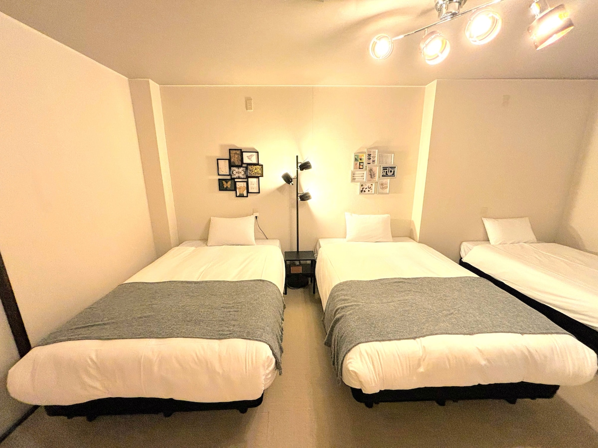 A spacious room features three comfortable beds, each covered with soft gray bedding. The arrangement allows for easy movement between the beds. Wall-mounted lights provide illumination, while framed photos and artwork enhance the decor, offering a cozy ambiance.