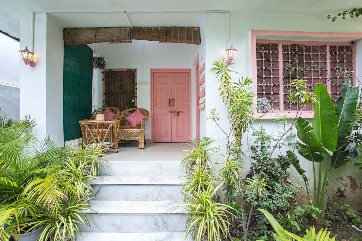 A vibrant entrance adorned with lush greenery and pastel accents invites guests into a serene atmosphere. Two wicker chairs are positioned near the pink door under warm lighting, and colorful plants frame the steps that lead up to the inviting entrance.