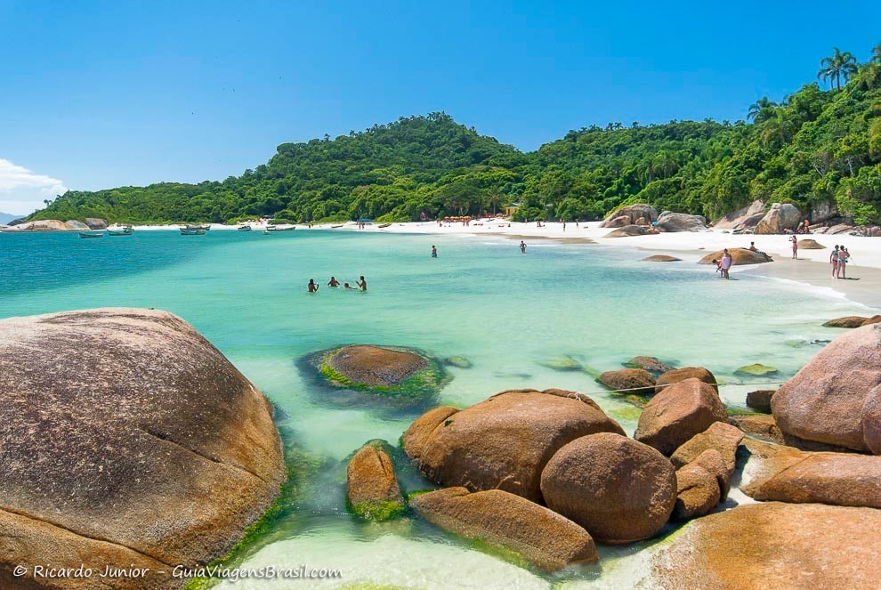 A tranquil beach scene is depicted, featuring clear turquoise waters gently lapping at smooth, rounded boulders. Lush green hills rise in the background, while individuals enjoy the coastal environment. The sandy shore is visible, with some visitors wading and relaxing along the water's edge.