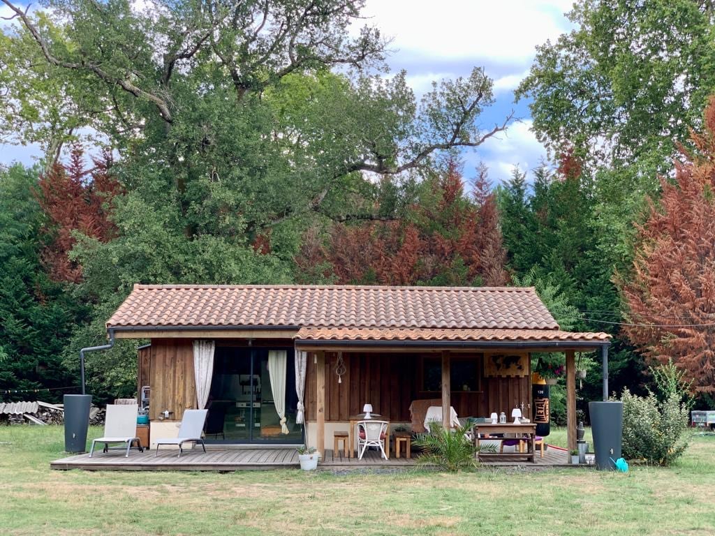 A wooden chalet features a sloping tiled roof and large windows, surrounded by greenery. A wooden deck with chairs and a table offers outdoor seating, creating a welcoming space to relax. The landscape includes a mix of trees in varying shades, adding to the natural setting.
