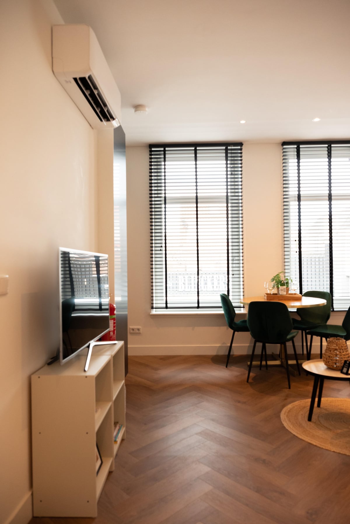 A modern living area is depicted, featuring a wall-mounted flatscreen TV positioned above a simple media unit. Large windows with black blinds allow natural light to fill the space, illuminating a dining table surrounded by green chairs. The flooring consists of herringbone-patterned wood.