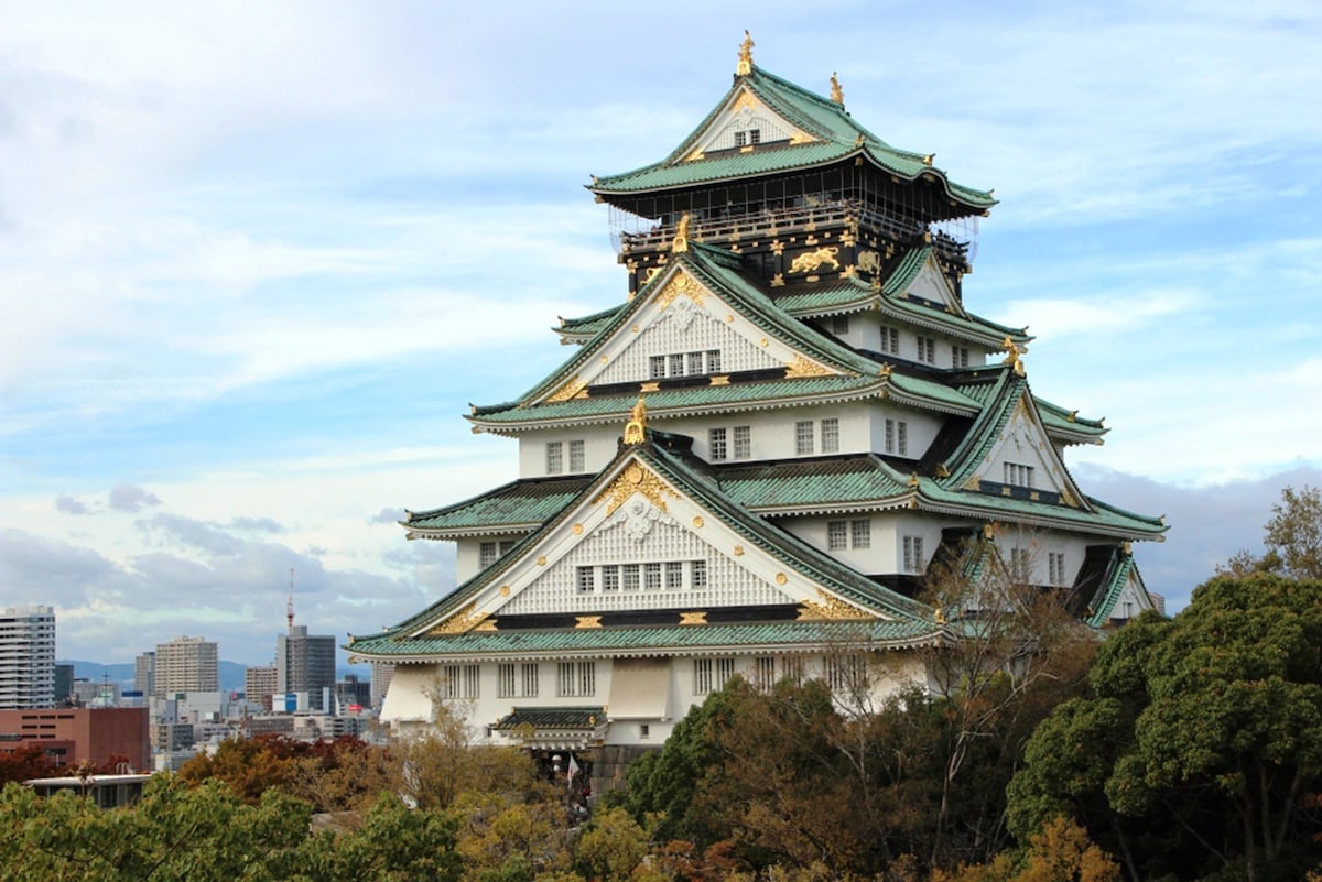 The iconic Osaka Castle is depicted with its distinctive green and gold roofs, framed by surrounding trees. The structure stands prominently against a clear sky, showcasing its traditional architectural features, while modern buildings can be seen in the background.