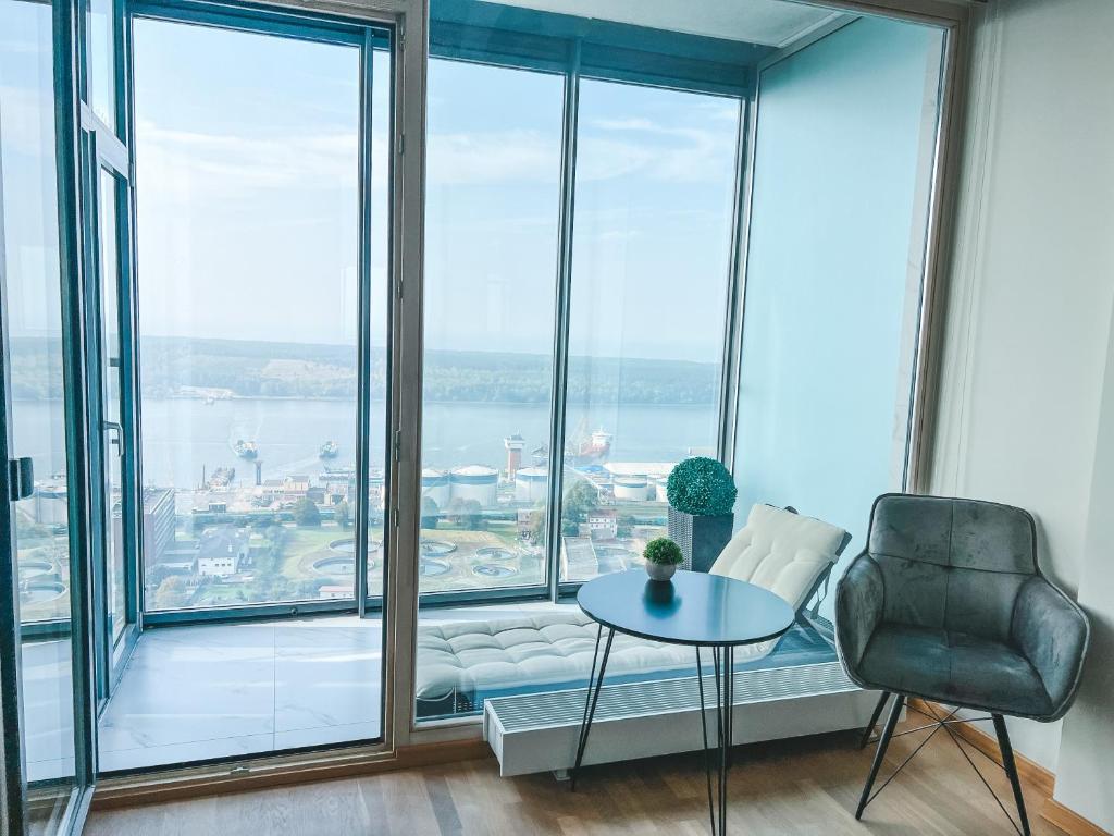 A spacious seating area is showcased with panoramic floor-to-ceiling glass windows, providing views of the sea and lagoon. A modern chair is positioned next to a minimalist round table, while a small potted plant adds a touch of greenery to the setting.