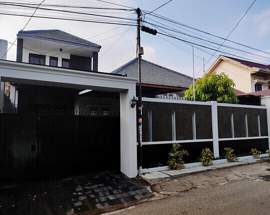 A two-story villa is displayed behind a secure black fence, providing privacy. The exterior features a well-maintained facade with a combination of dark and light tones. Lush green plants are seen along the fence, contributing to the inviting entrance.