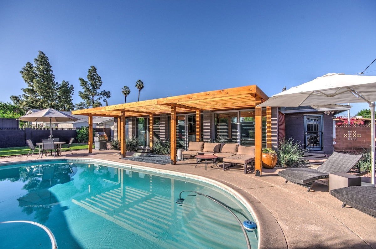 A charming outdoor space features a sparkling swimming pool surrounded by a spacious patio. Sleek lounge chairs and shaded areas offer relaxation options. A wooden pergola provides structure while enhancing the connection between the home and garden, with lush greenery in the background.