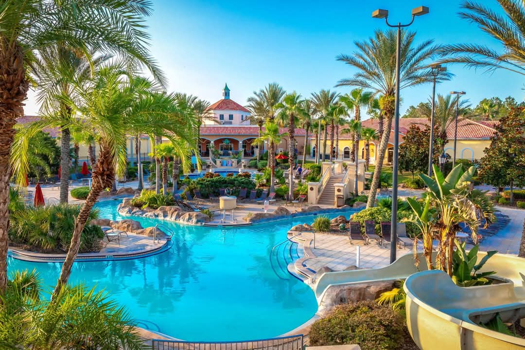 A vibrant tropical resort features a sprawling pool complex surrounded by palm trees. The clear blue water reflects sunlight, with winding pathways leading to several water features, including a water slide and lazy river. Colorful loungers and umbrellas are positioned throughout the inviting outdoor space.