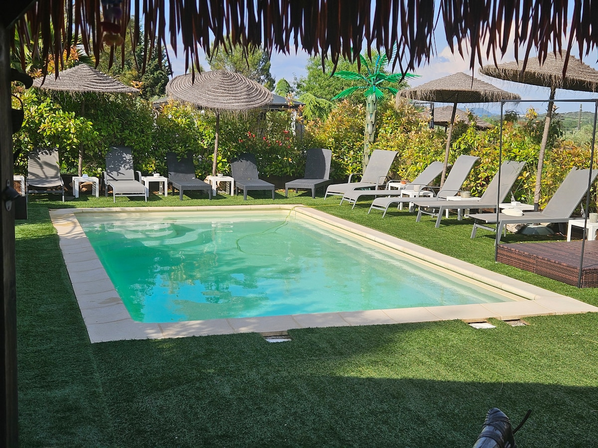 A private pool area is surrounded by landscaped greenery and umbrellas for shade. Chaise lounges are neatly arranged around the pool, providing a comfortable space for relaxation. The clear water of the pool reflects the sunlight, creating a welcoming atmosphere.