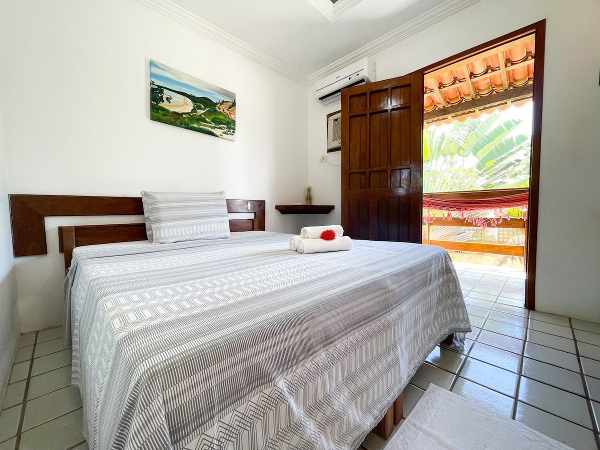 A comfortable sleeping area features a spacious bed dressed in a striped blanket. A wooden door leads to a balcony, revealing outdoor greenery. Decorative wall art and air conditioning enhance the room's comfort, while tiled floors provide a clean and simple aesthetic.