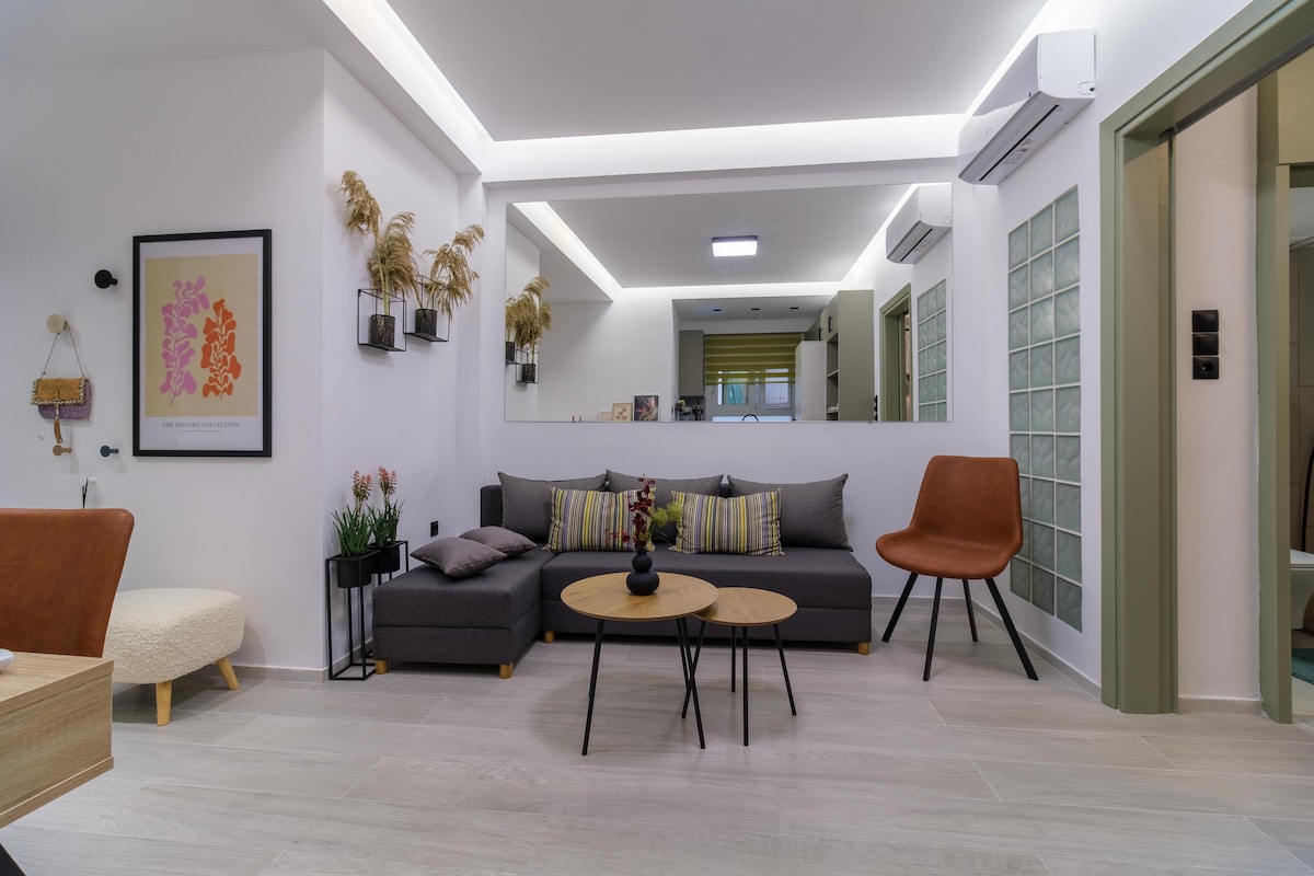 The living area features a modern seating arrangement with a pull-out couch adorned with colorful cushions. Two round coffee tables are positioned at the center, surrounded by stylish chairs. A large mirror reflects the space, enhancing brightness, while minimal artwork adds character to the wall.