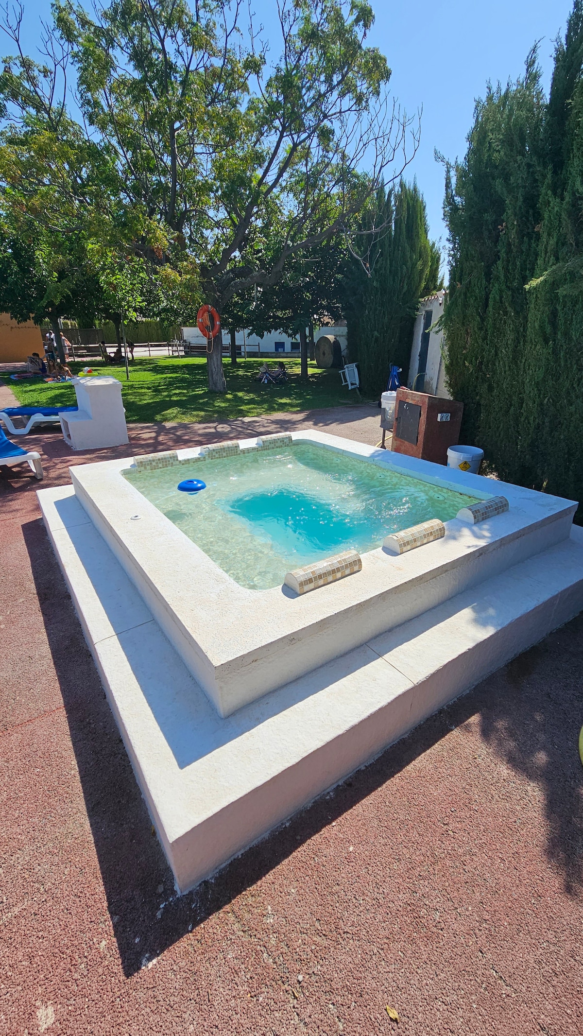 A jacuzzi is set within a spacious outdoor area, surrounded by greenery and a paved surface. The water is clear and inviting, while steps lead up to the jacuzzi's platform. Several lounge chairs can be seen nearby, contributing to a relaxed atmosphere.