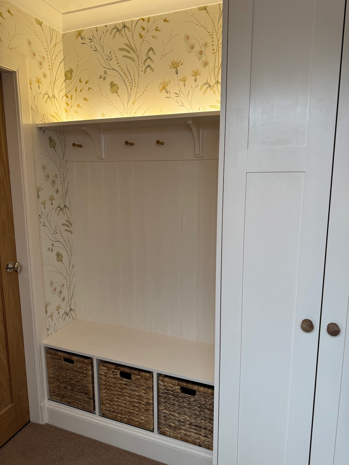 A built-in entryway nook is featured, with a light-colored shelf above for storage. Three woven baskets are neatly arranged below, offering organization. The area is complemented by floral-patterned wallpaper that adds a touch of nature to the space.