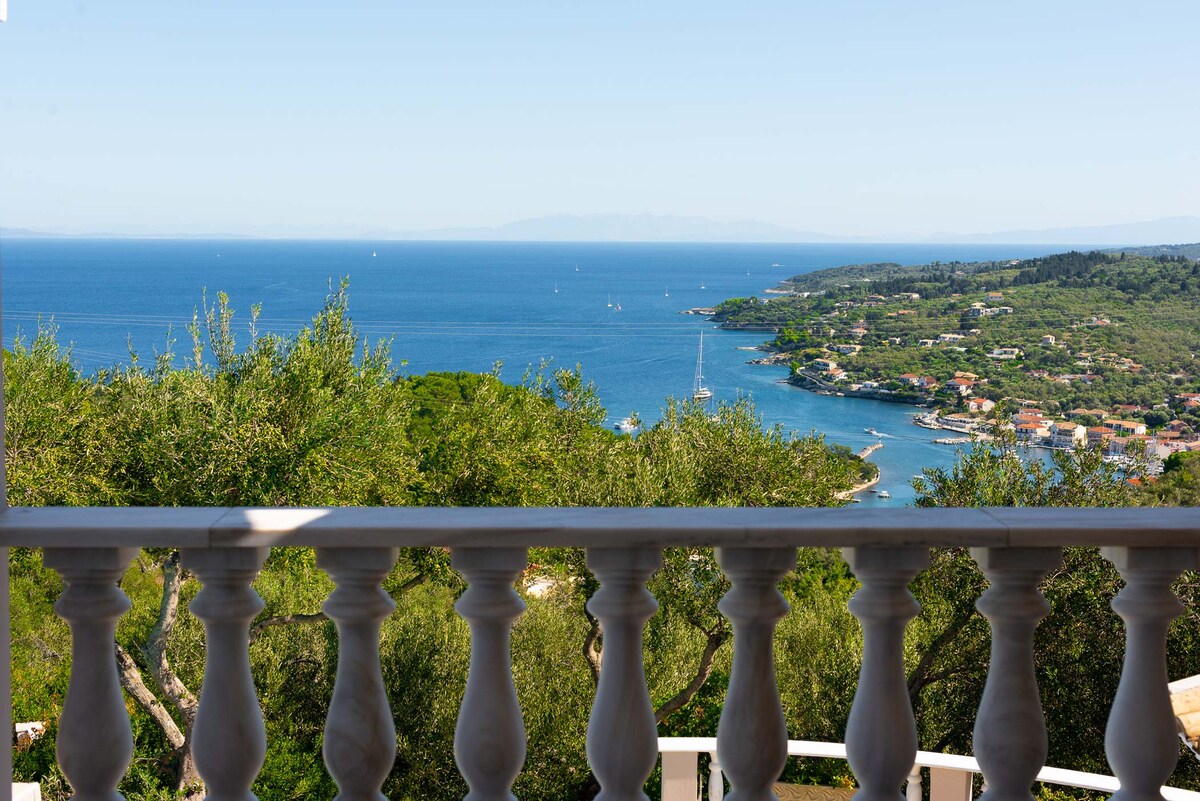 A scenic view is presented from a terrace railing, overlooking the coastline and shimmering sea. Lush green hills and colorful houses line the shore, while boats can be seen dotting the tranquil waters under a clear blue sky.