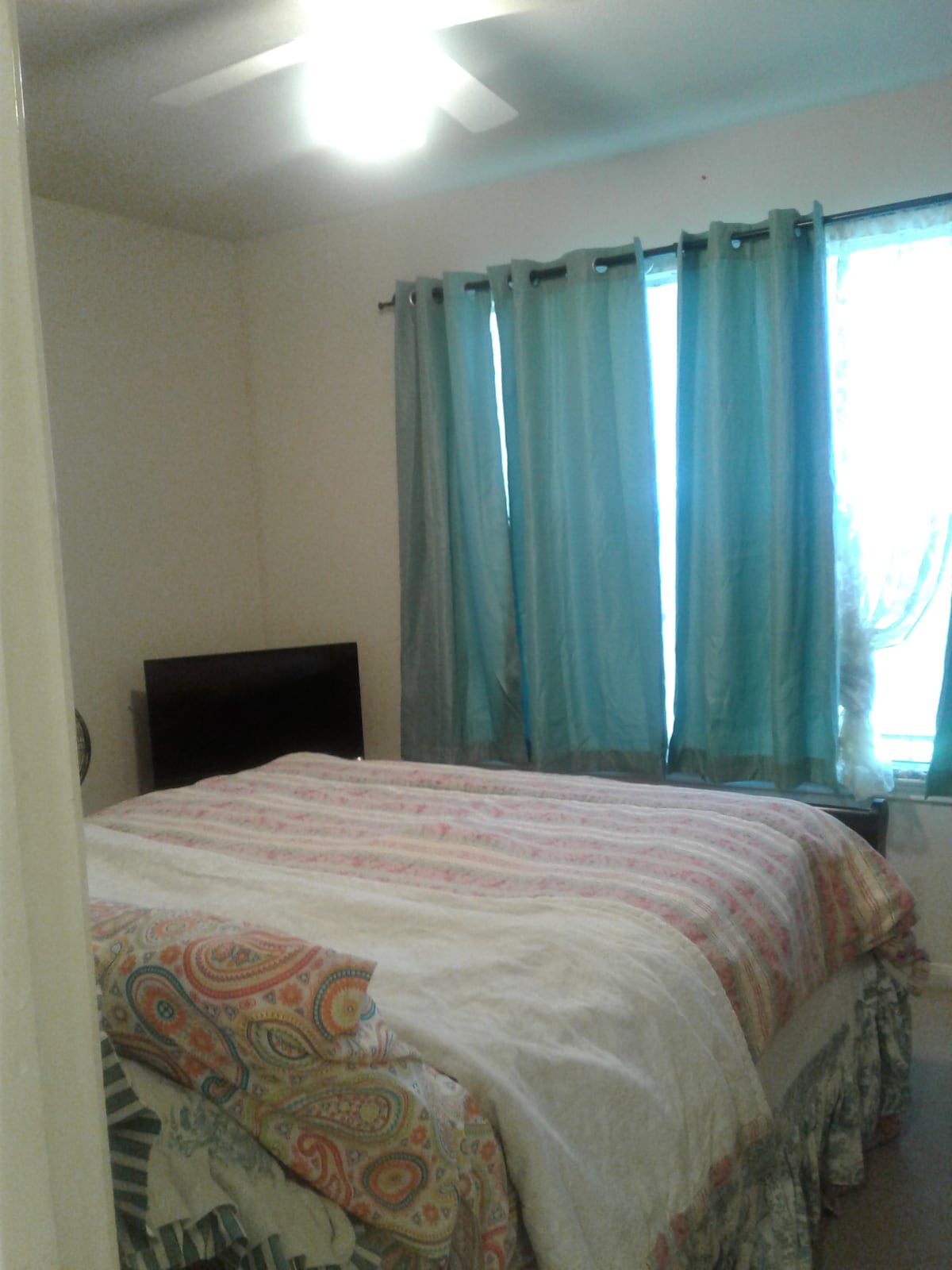 A spacious room features a queen size bed with a patterned comforter. A dresser is positioned against the wall, and ceiling fan is visible above. Light filters through large windows dressed in light, airy curtains, creating a calming atmosphere.