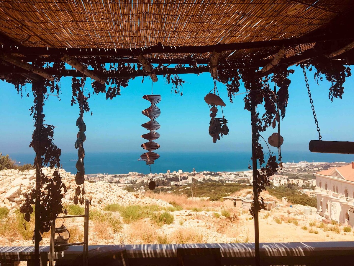 A shaded outdoor seating area is framed by a straw canopy, adorned with hanging decorations. In the background, expansive views of the sparkling blue sea and the distant city skyline are visible, complemented by the greenery of the surrounding hills.
