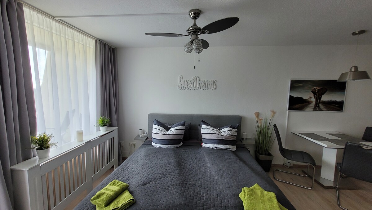 A spacious bedroom is presented, featuring a large bed with dark linens and decorative pillows. Natural light filters through sheer curtains, illuminating the soothing grey walls. A small table and chairs are arranged nearby, with potted plants adding a touch of greenery.