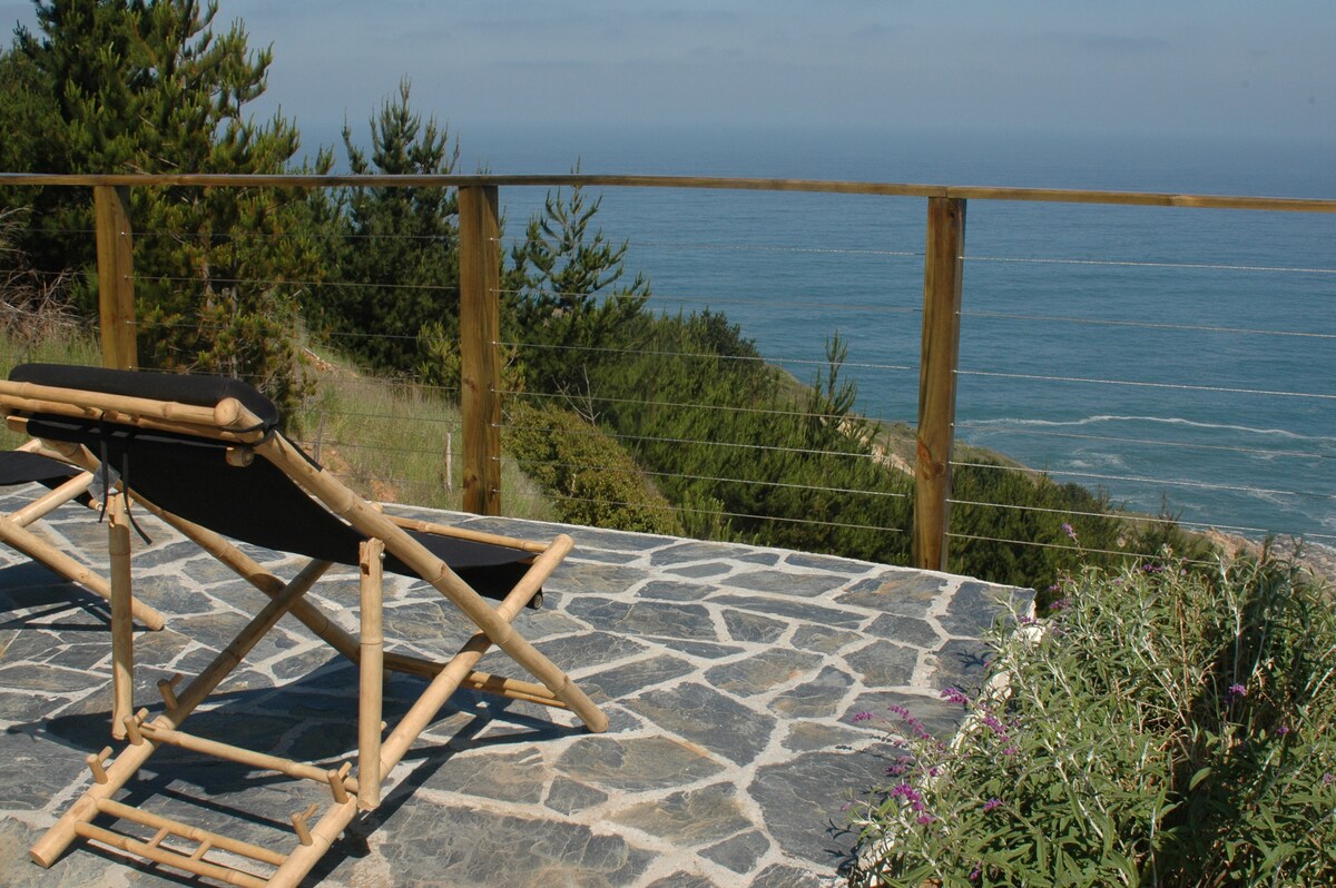 A wooden terrace features a reclining chair positioned to overlook the ocean, providing views of the water and surrounding greenery. The terrace is bordered by a wooden railing, and the stone flooring complements the natural setting.