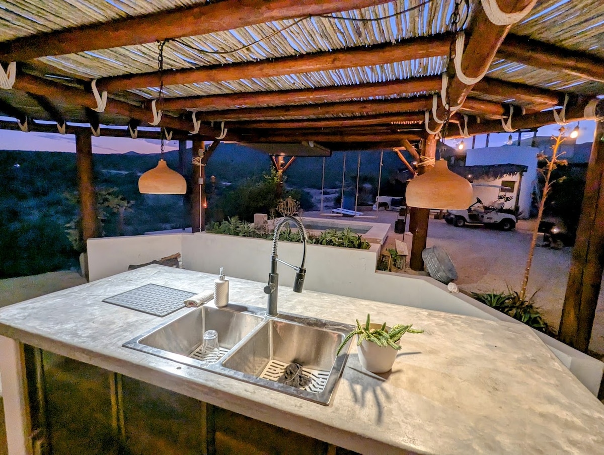 An outdoor kitchen area is featured under a wooden pergola, with a spacious concrete countertop and double sink. Soft lighting is provided by hanging fixtures. Surrounding greenery adds a natural touch, while a glimpse of the desert landscape can be seen in the background.