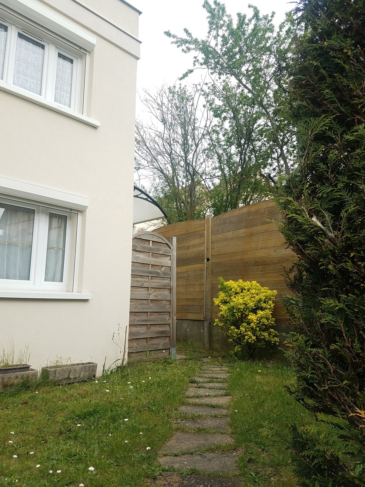 A grassy pathway leads through a quaint garden area, bordered by wooden fencing and soft greenery. A flowering bush adds a touch of color, while trees provide a natural backdrop. The space appears serene, providing a quiet retreat just outside the home.