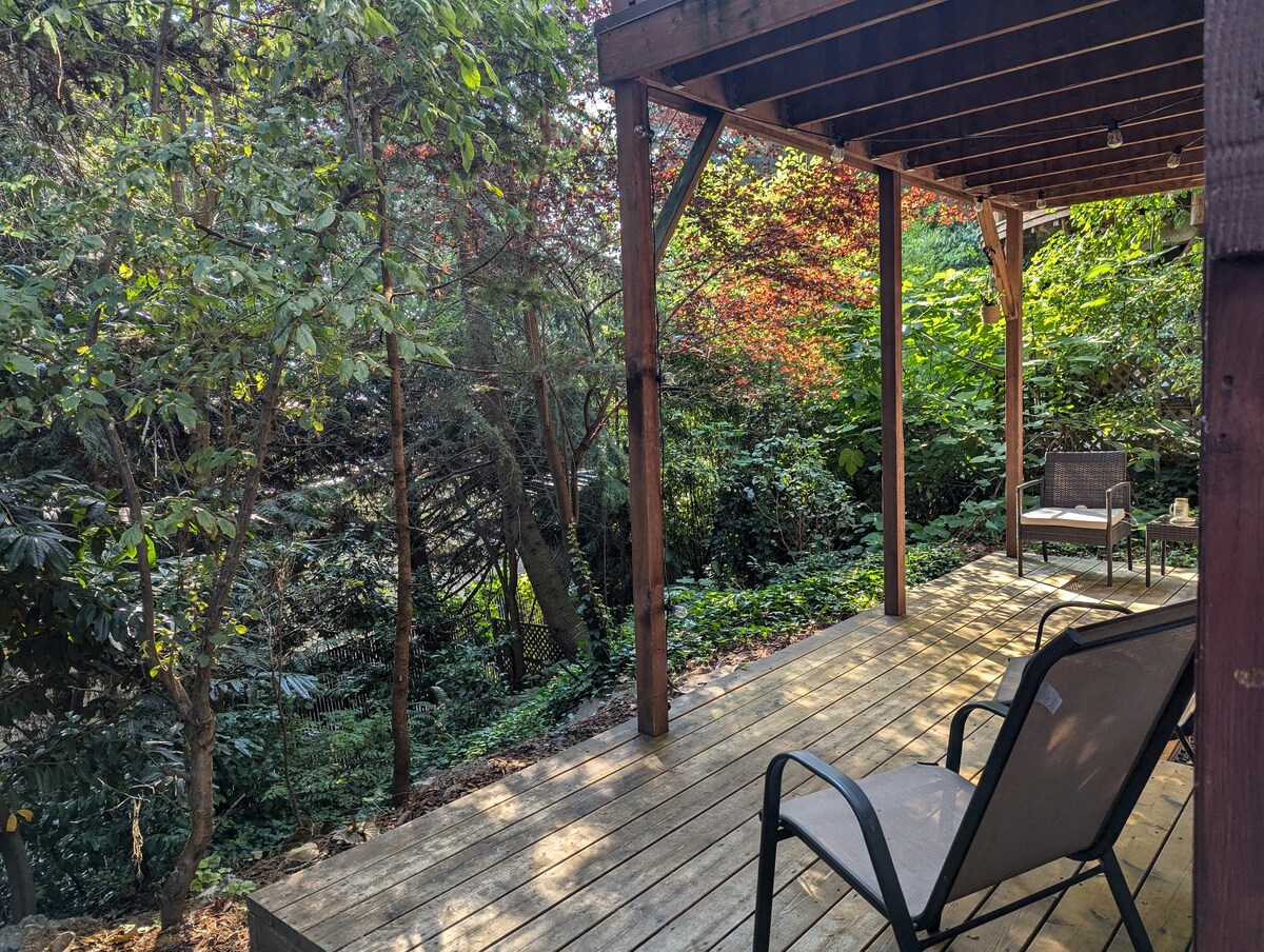 A private porch is depicted, featuring wooden decking and a covered area supported by beams. Two chairs are positioned to the side, overlooking a lush green landscape filled with trees and foliage, providing a serene outdoor space.