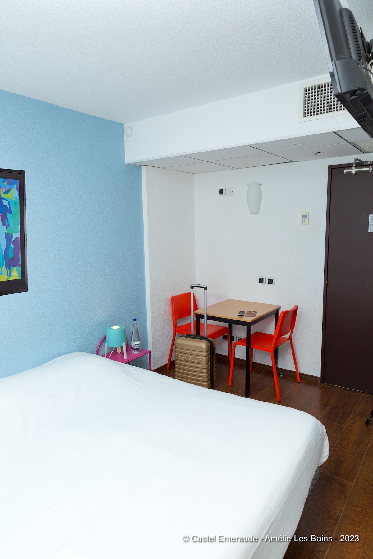 A double room features a spacious bed covered in crisp white linens. A small table is paired with two bright red chairs. A luggage piece is placed beside the bed, and a water bottle is positioned on the side table against a blue wall.