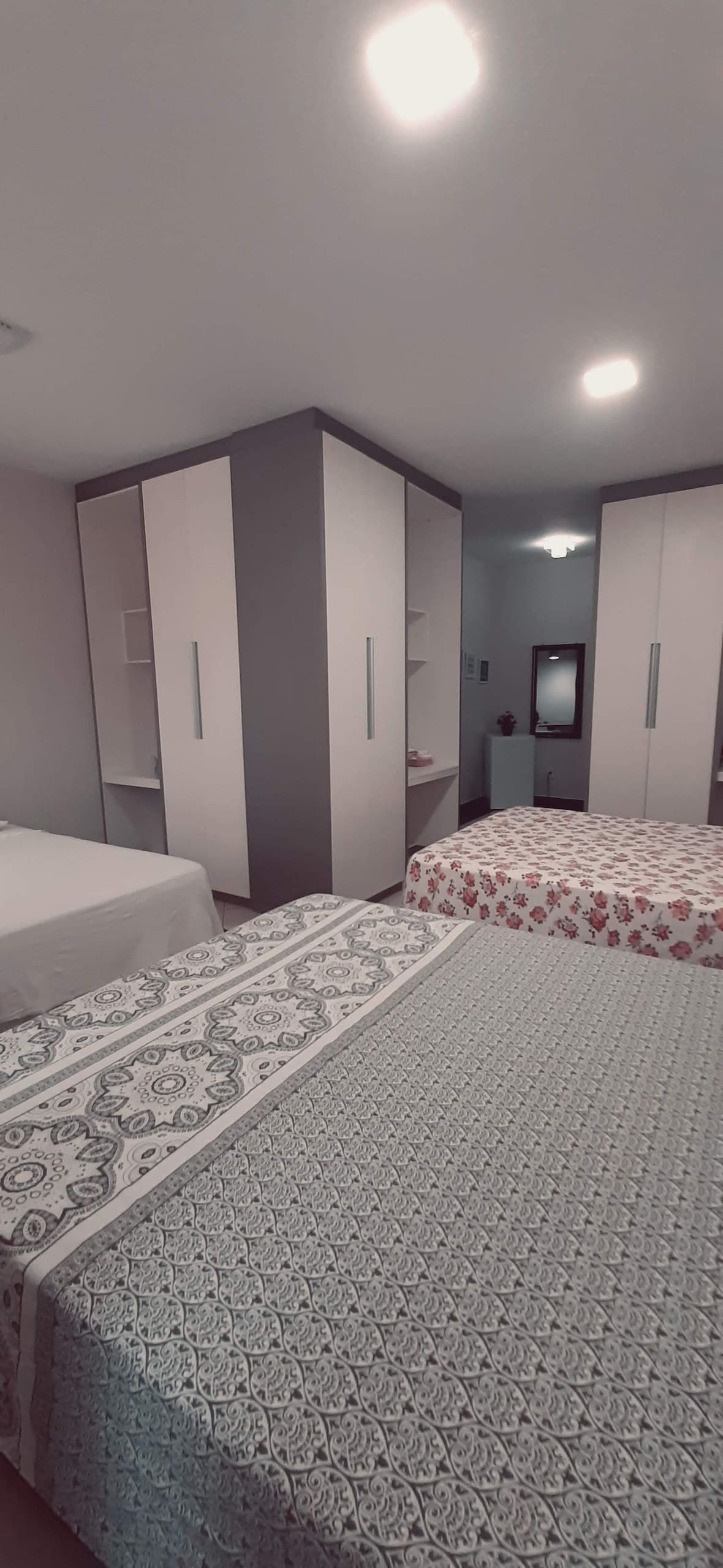 A spacious bedroom is shown, featuring three queen-size beds with diverse patterned bed linens. Individual wardrobes are positioned along the walls, and a small mirror is visible in the background. Soft lighting enhances the room's inviting environment.