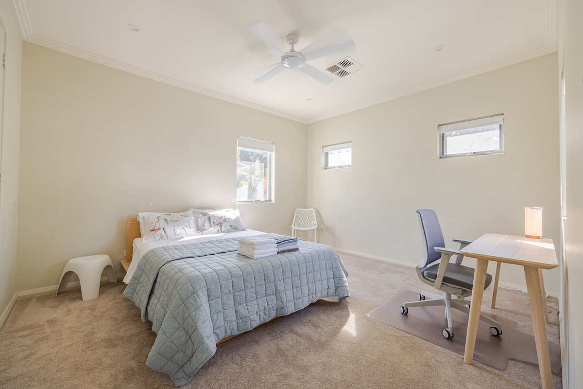 A spacious bedroom features a large bed with a light-colored quilt, accented by decorative pillows. A comfortable desk and chair are positioned beside a lamp, while two small windows allow natural light to fill the room. Soft carpeting covers the floor.