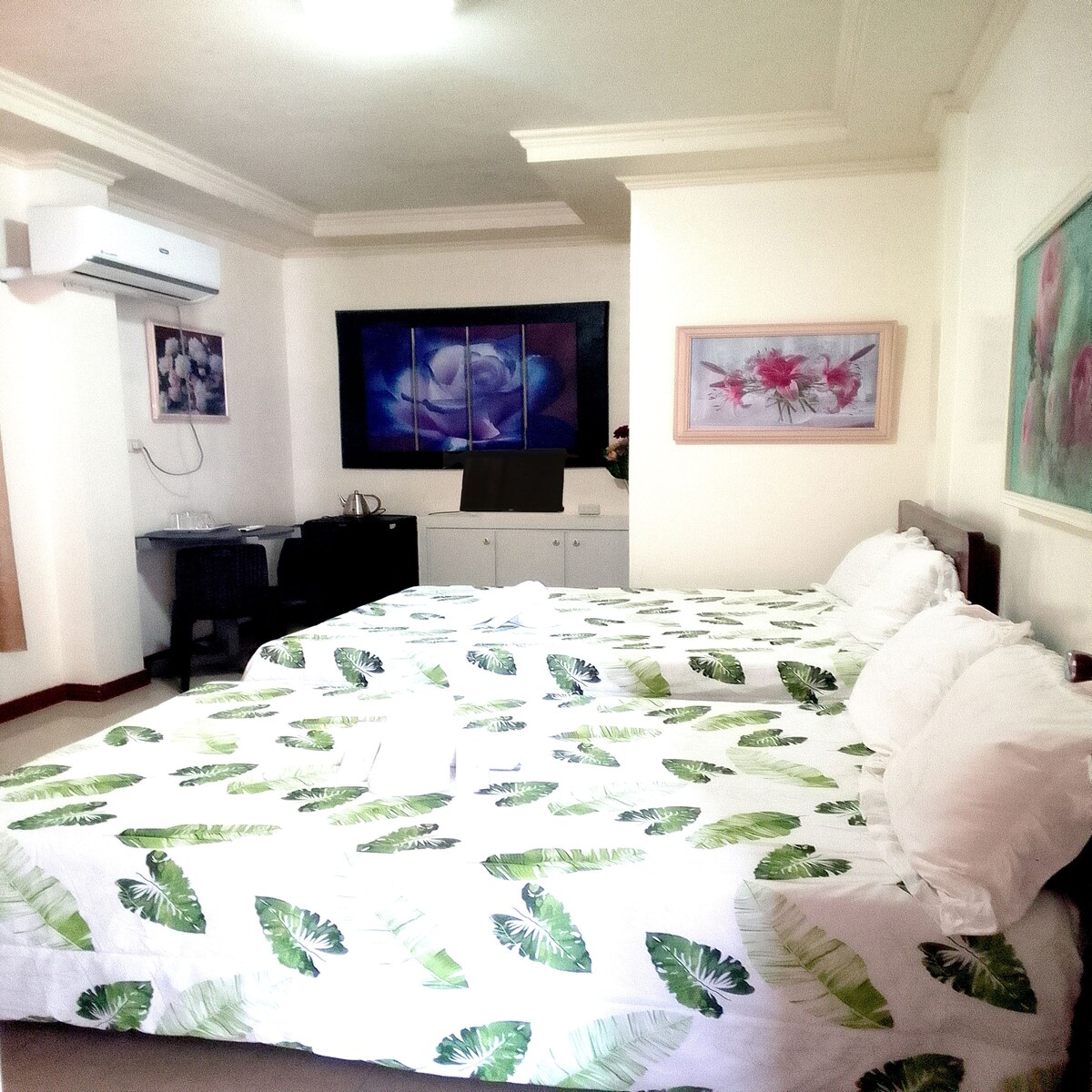 A spacious bedroom is furnished with two queen size beds, dressed in leaf-patterned linens. Artwork featuring floral designs adorns the walls, and a desk setup is visible in the background. Natural light enters from a window, contributing to a bright ambiance.