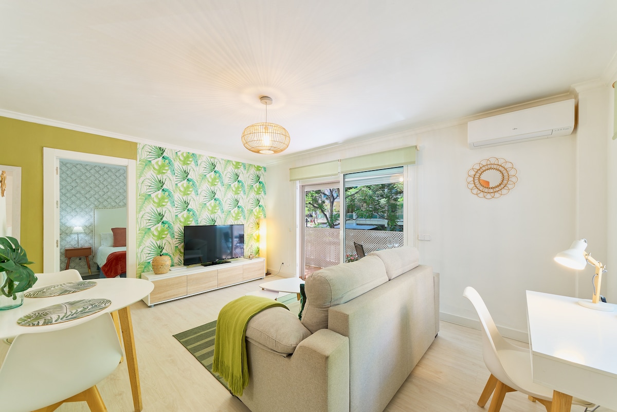 A stylish living room is presented, featuring a comfortable sofa and a sleek TV unit. A dining table with chairs occupies one corner, and a bright workspace is set up alongside. Lush green wallpaper and natural light from the terrace enhance the inviting ambiance.