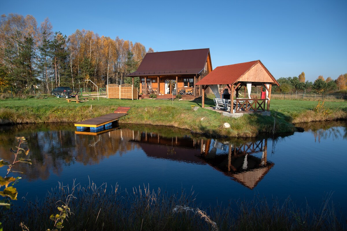 A charming wooden house is reflected in a serene pond, surrounded by lush greenery. Adjacent to the house, an open gazebo offers shaded seating and leisure space. The peaceful landscape is enhanced by trees displaying autumn foliage under a clear blue sky.
