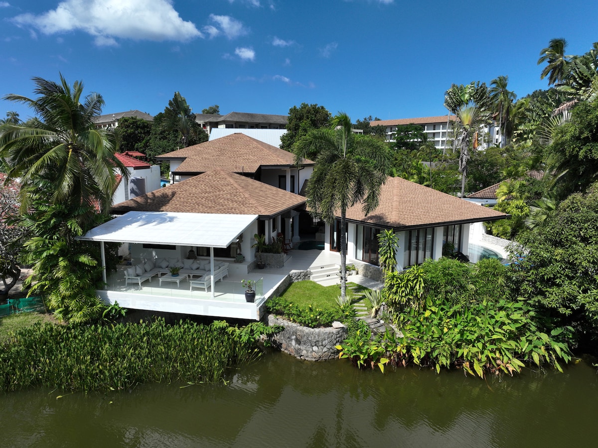 A spacious villa is set along a lush green landscape, bordered by a tranquil lake. The structure features a modern design with a sloping roof and expansive outdoor seating areas. Surrounding tropical vegetation adds to the serene ambiance, creating a peaceful retreat.