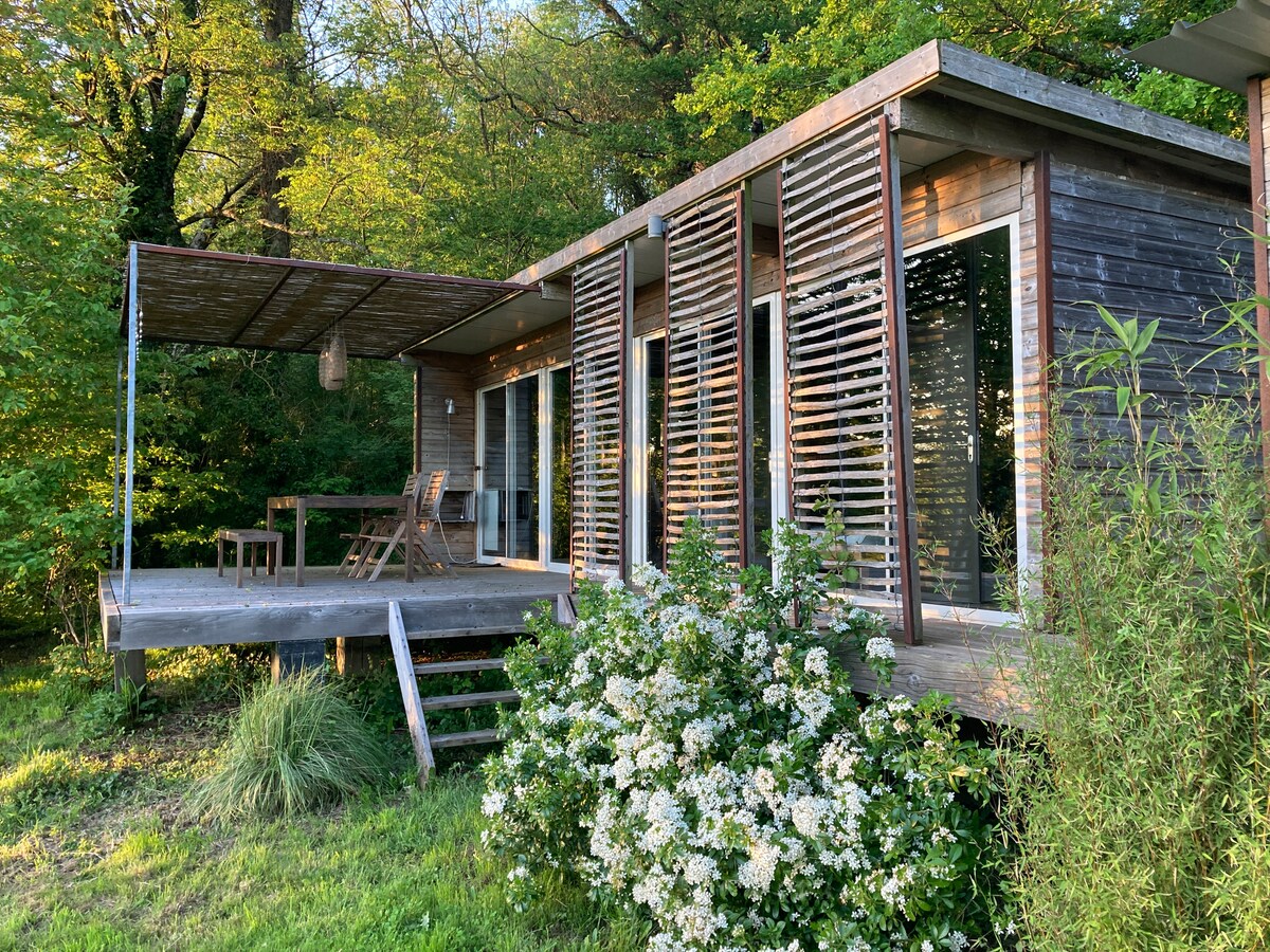 The wooden lodge is nestled amid lush greenery, featuring large glass doors that open onto a spacious terrace. A small dining table and chairs are visible on the deck. Flowering bushes and grass surround the exterior, providing a natural and serene setting.