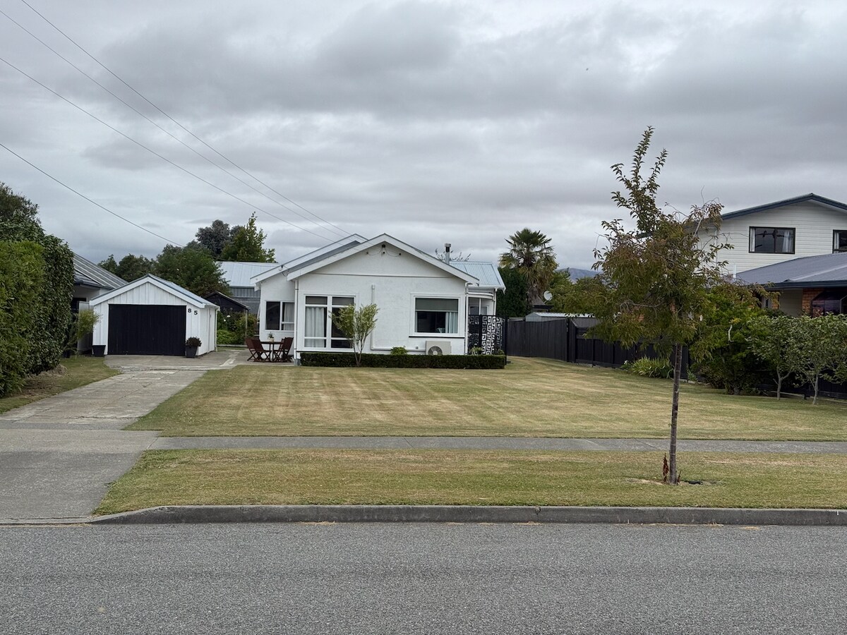 A single-story white cottage is set back from the road, surrounded by a well-maintained lawn. A driveway leads to a garage, while a patio with outdoor furniture is visible in front. Neatly trimmed hedges frame the property, enhancing its curb appeal.