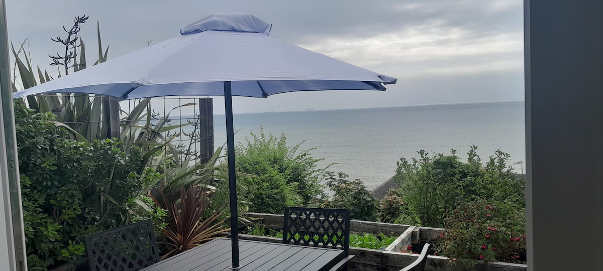 A dining area is set on a decked terrace, featuring a table and black chairs beneath a large, light blue umbrella. Lush green plants surround the space, with a view of the sea under an overcast sky in the background.