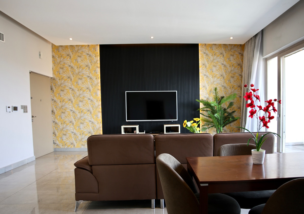 A bright living area is showcased, featuring a flat-screen TV mounted on a textured black wall. A dark brown sofa offers comfortable seating, complemented by a wooden dining table. Large glass doors lead to an outdoor space, while decorative plants add a touch of greenery.