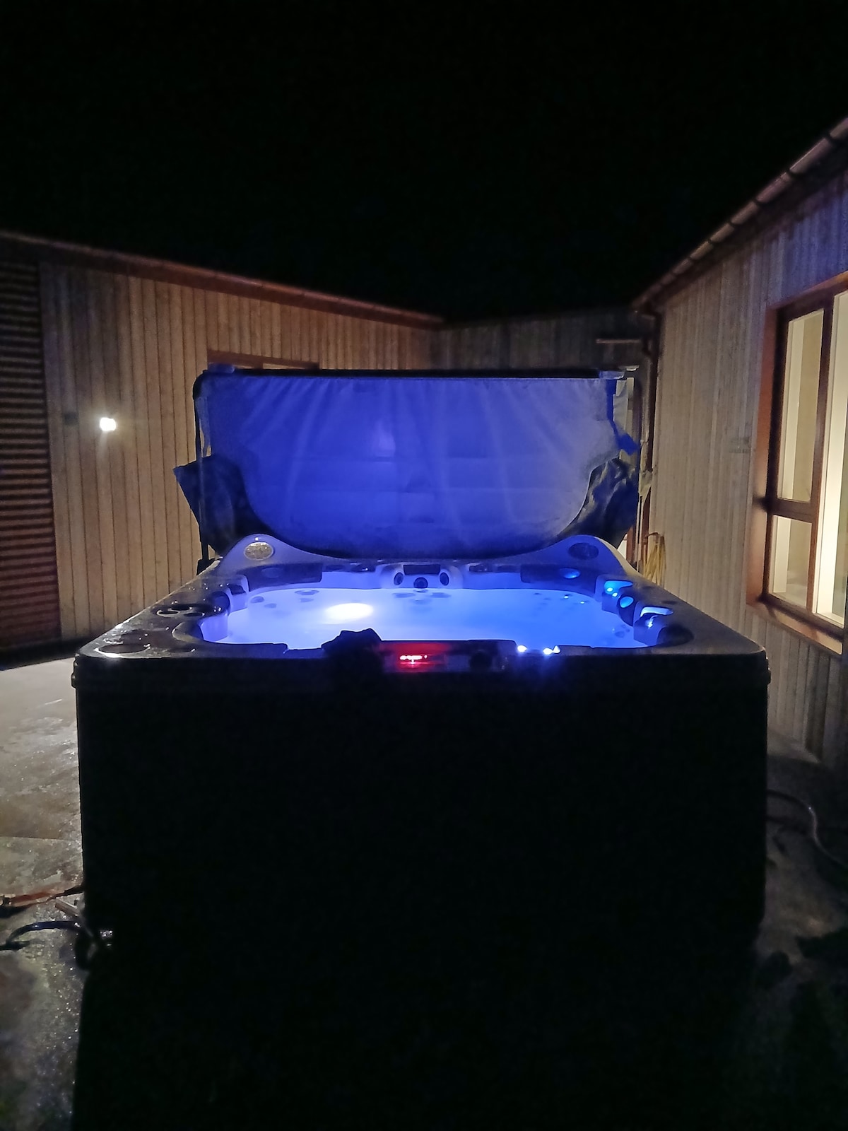 An outdoor hot tub is illuminated with soft blue lights, creating a calming atmosphere in the night. The hot tub's cover is lifted, revealing the inviting interior. Nearby, a window hints at a cozy accommodation, adding to the tranquil setting.