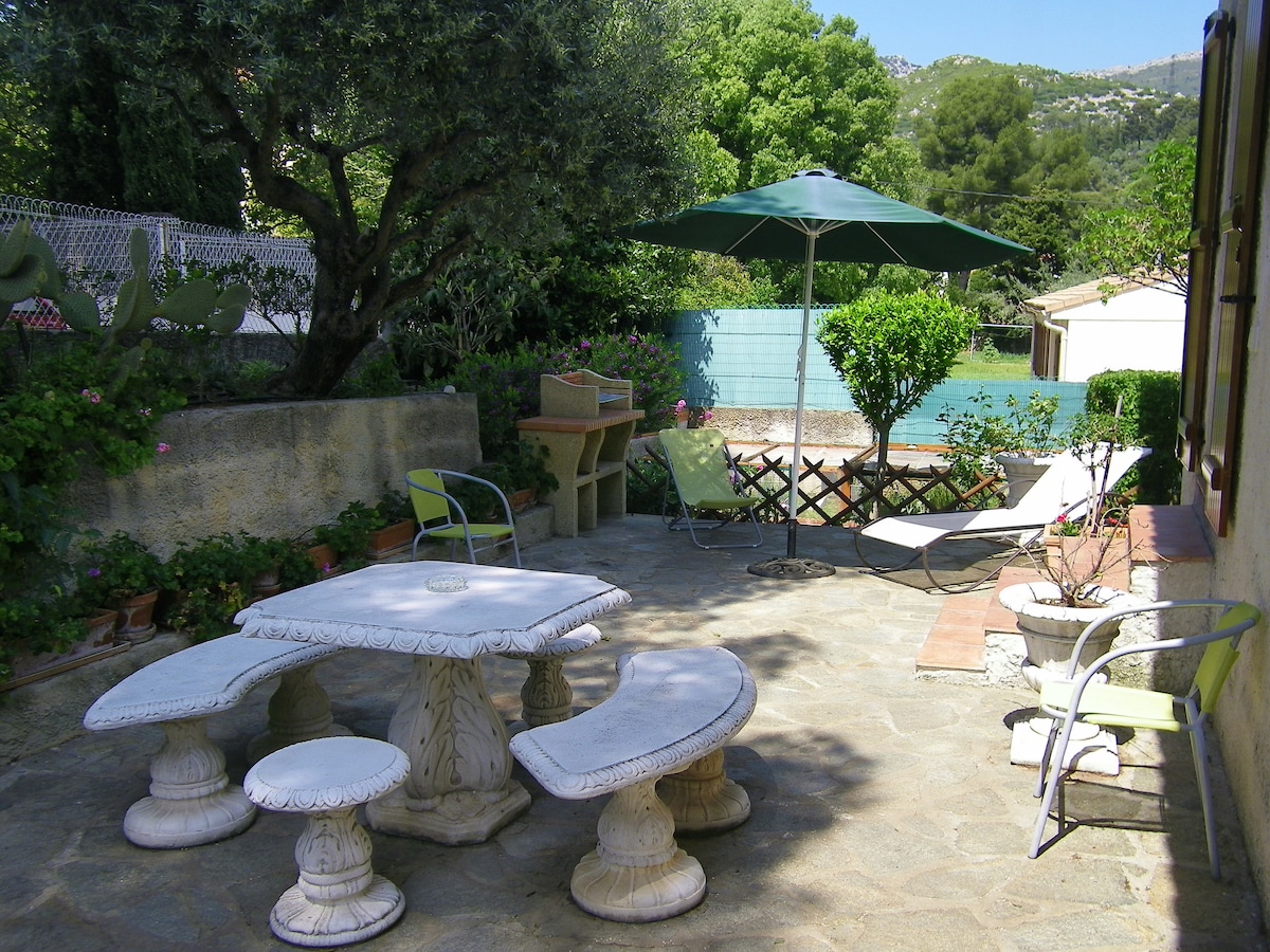 A private outdoor space is identified, featuring a stone-paved terrace surrounded by greenery. A circular table with matching chairs occupies the center, alongside a large umbrella for shade. Additional seating is provided by a lounge chair, while a barbecue stands ready for outdoor cooking.