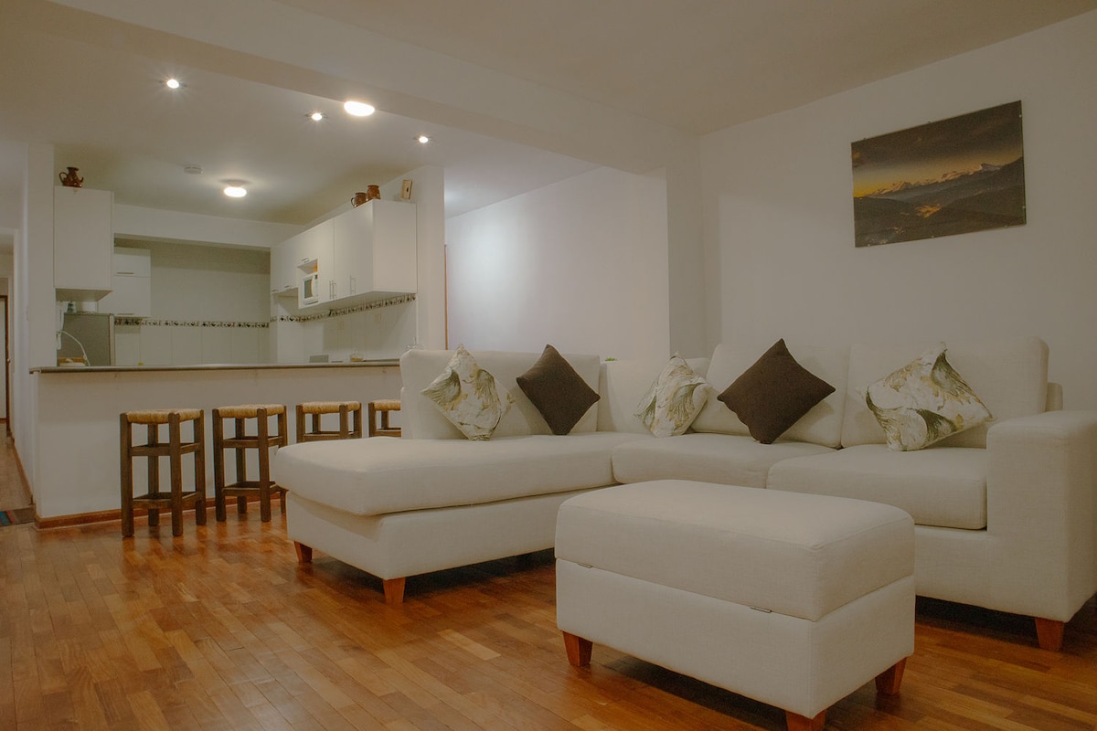 A light-filled living area features a spacious sectional sofa adorned with patterned cushions. A small ottoman complements the seating. In the background, a kitchen area with white cabinetry is visible, along with four wooden stools positioned at a low counter.