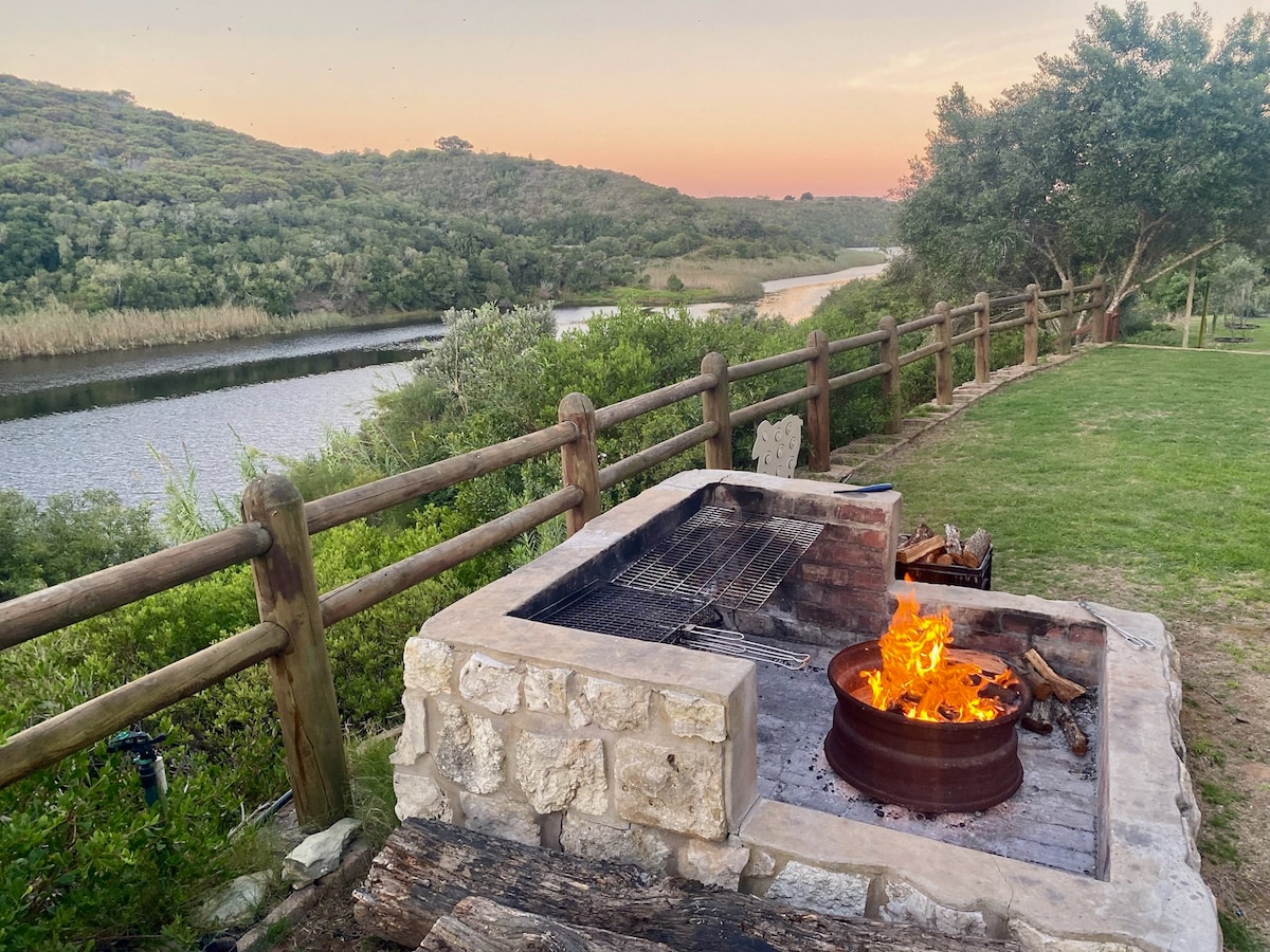 An outdoor firepit is situated near the riverbank, surrounded by a grassy area and wooden fencing. Flickering flames generate warmth as the sun sets over the serene landscape, with lush greenery lining the river and rolling hills in the background.