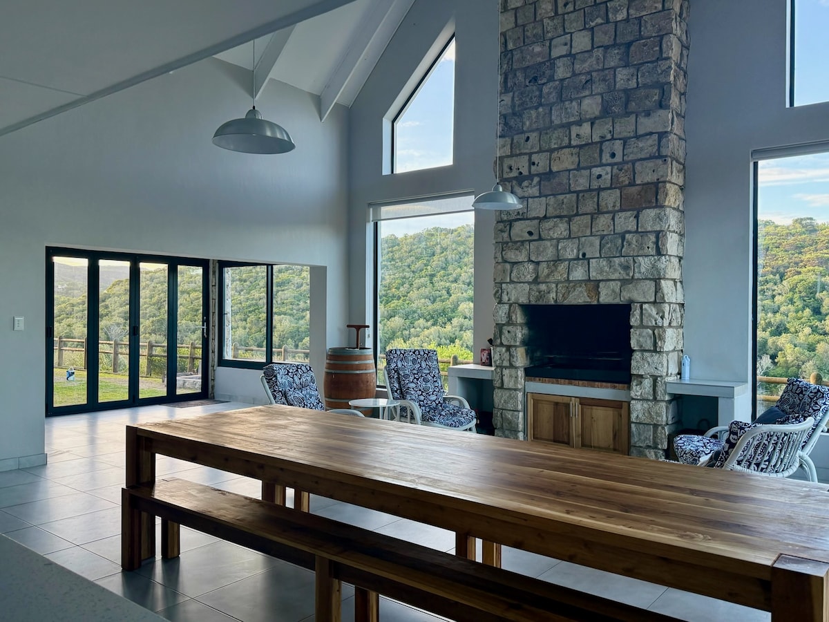 A spacious living area features large glass doors offering views of the surrounding hills. A rustic stone fireplace serves as a focal point. A wooden dining table occupies the center, while two comfortable chairs are positioned by the fireplace, creating an inviting gathering space.