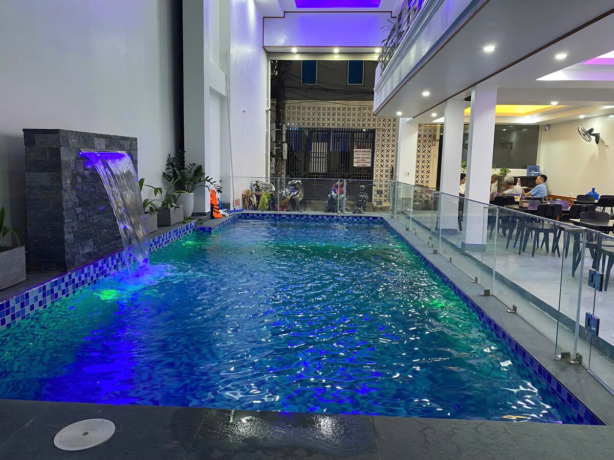 An indoor pool is visible, featuring gentle flowing water cascading from a stone structure. The pool's surface reflects a blend of blue and green lighting, enhancing the serene environment. Glass barriers surround the pool area, with seating and dining areas seen in the background.
