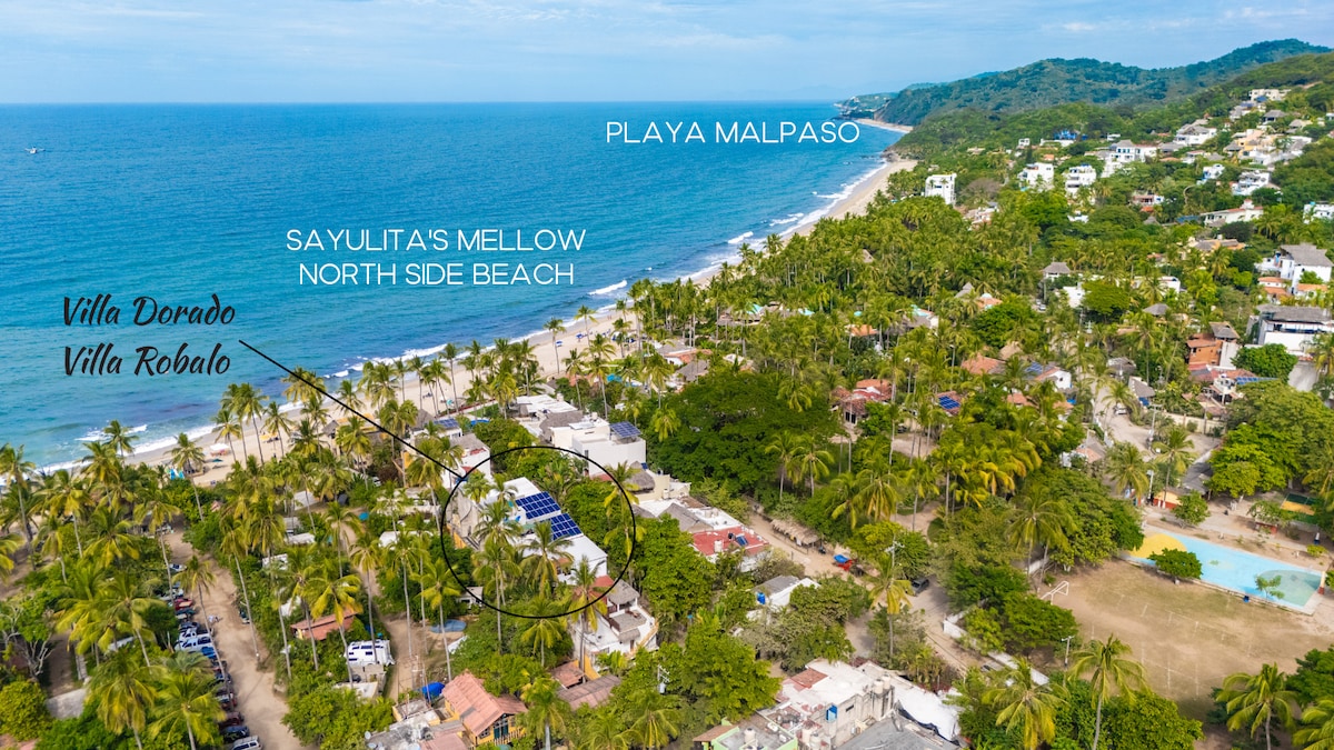 An aerial view captures the serene landscape of Sayulita's north side beach, with palm trees lining the shore. Villas Dorado and Robalo are positioned in the foreground, featuring a solar panel array. The coastline extends in the background, showcasing Playa Malpaso.