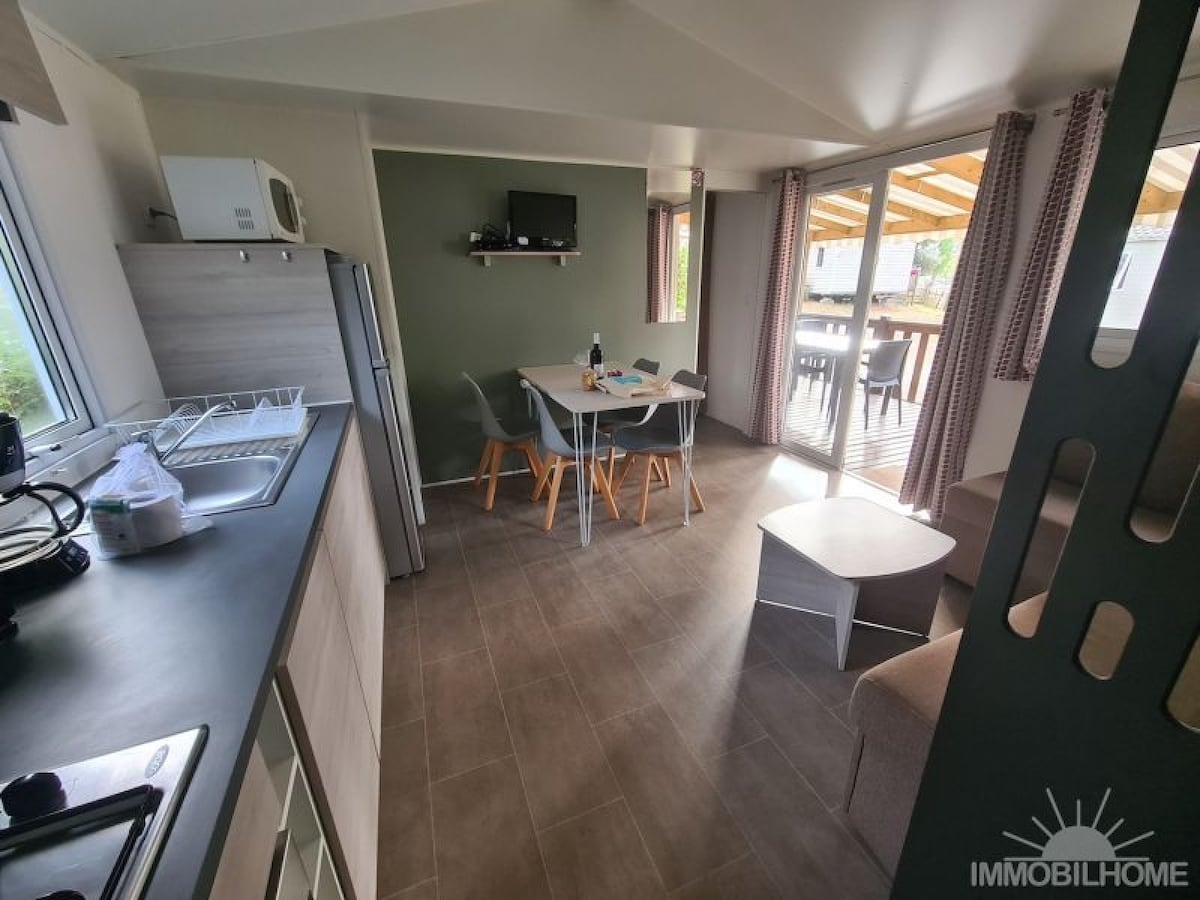 An open-plan living area features a modern kitchen with a refrigerator, a dining table with four chairs, and a cozy seating area. Bright natural light filters in through large windows, leading to an outdoor terrace with covered seating.