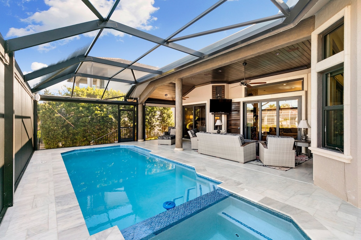 A covered outdoor area features a spacious swimming pool surrounded by marble flooring. Comfortable seating arrangements with light-colored furniture provide a relaxing atmosphere. Large glass doors offer views of the interior space, while lush greenery is visible through the screening.