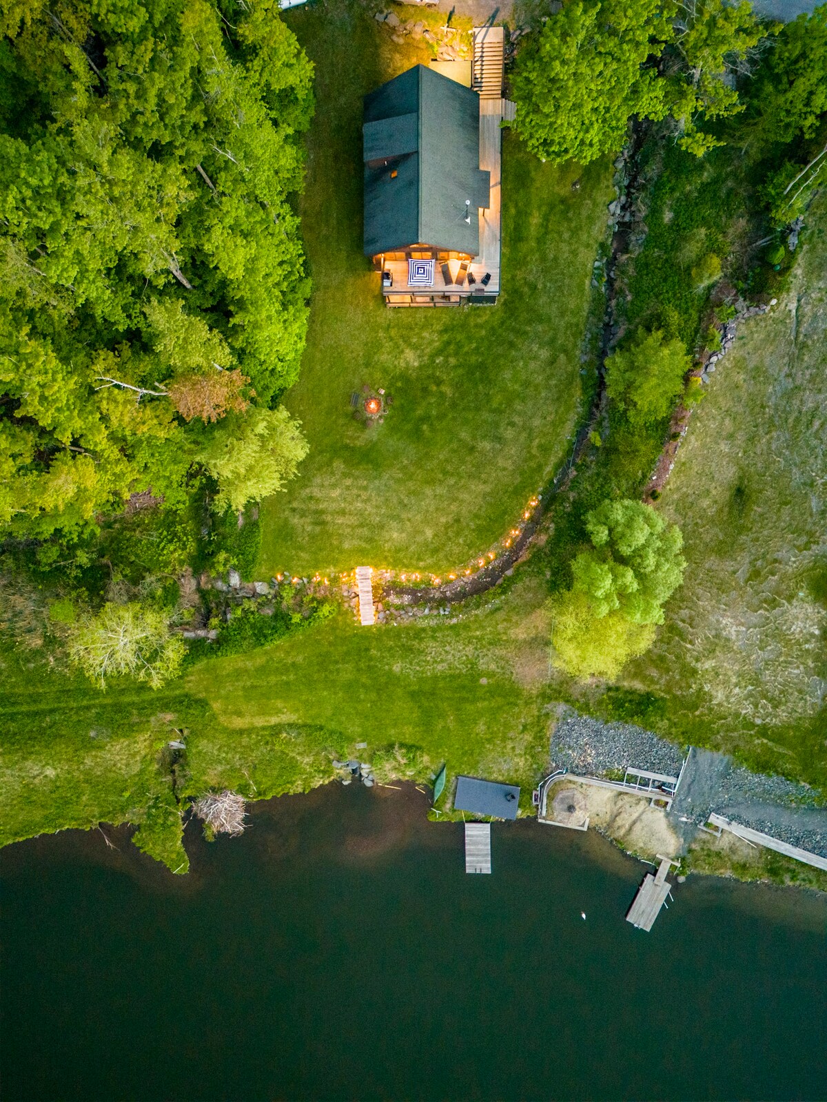 An aerial view showcases a house surrounded by lush greenery, alongside a tranquil lake. A well-maintained lawn leads to a private dock, where a small boat is visible. Pathway lights illuminate the winding path from the house to the lake, enhancing the serene setting.