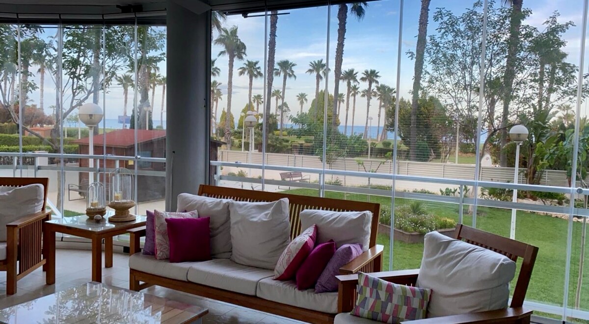 A spacious terrace is furnished with comfortable seating, featuring light-colored cushions accented with vibrant pillows. The area is surrounded by large glass panels, offering unobstructed views of palm trees and the ocean beyond, creating a serene outdoor retreat.
