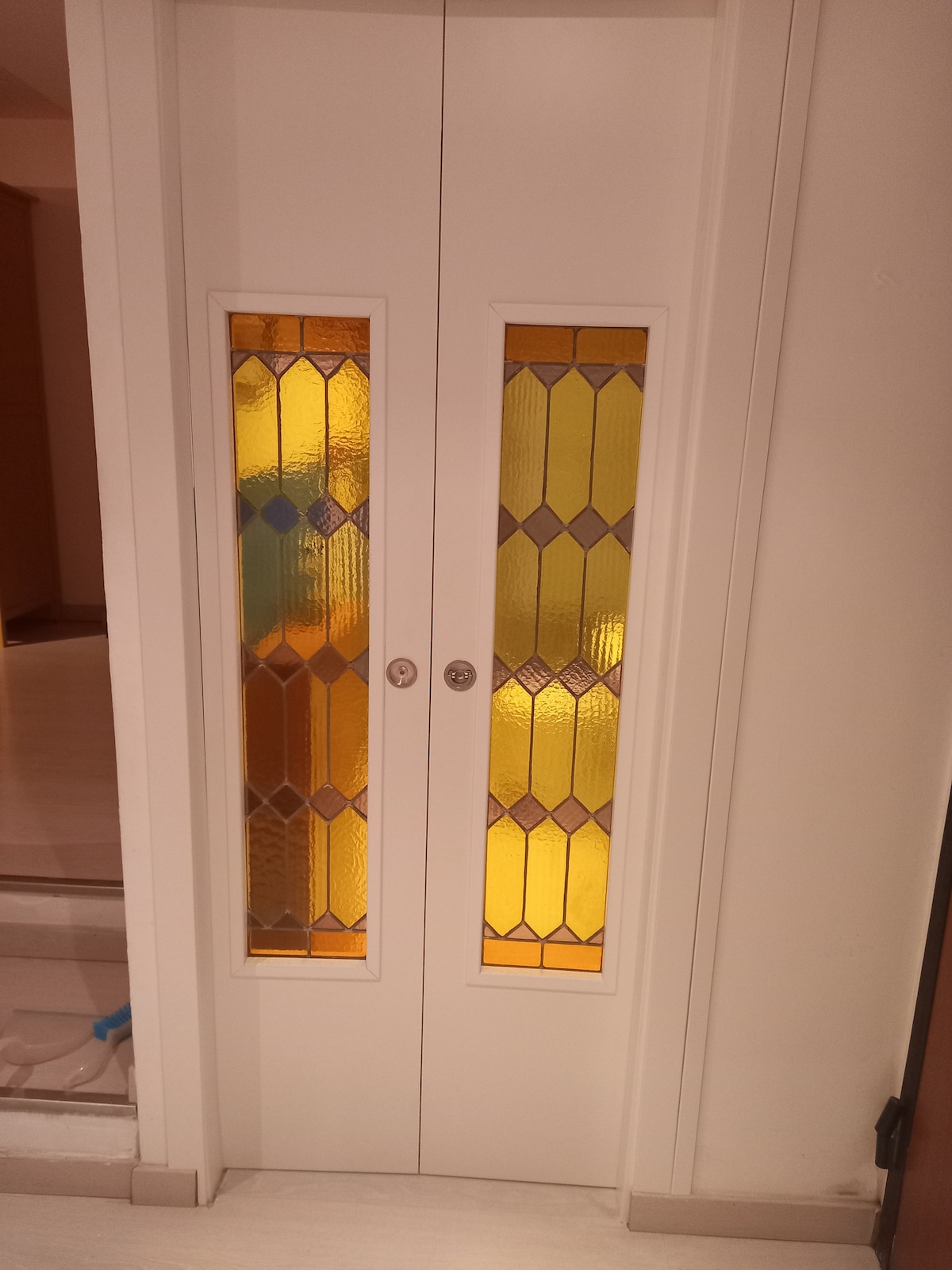 A double door is framed in white, featuring tall stained glass panels with warm tones of yellow and orange. The glass design incorporates geometric patterns, providing a blend of privacy and light to the interior space.