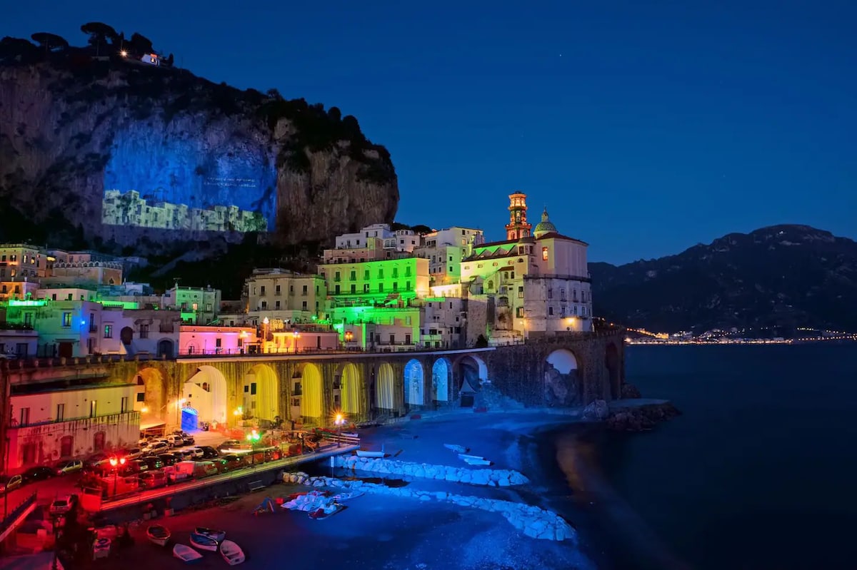 The coastal town of Atrani is illuminated at night, showcasing colorful lights reflecting off the buildings and water. The image captures the charming architecture along the shoreline and the cliffs surrounding the area, creating a serene atmosphere that highlights the beauty of the Amalfi Coast.